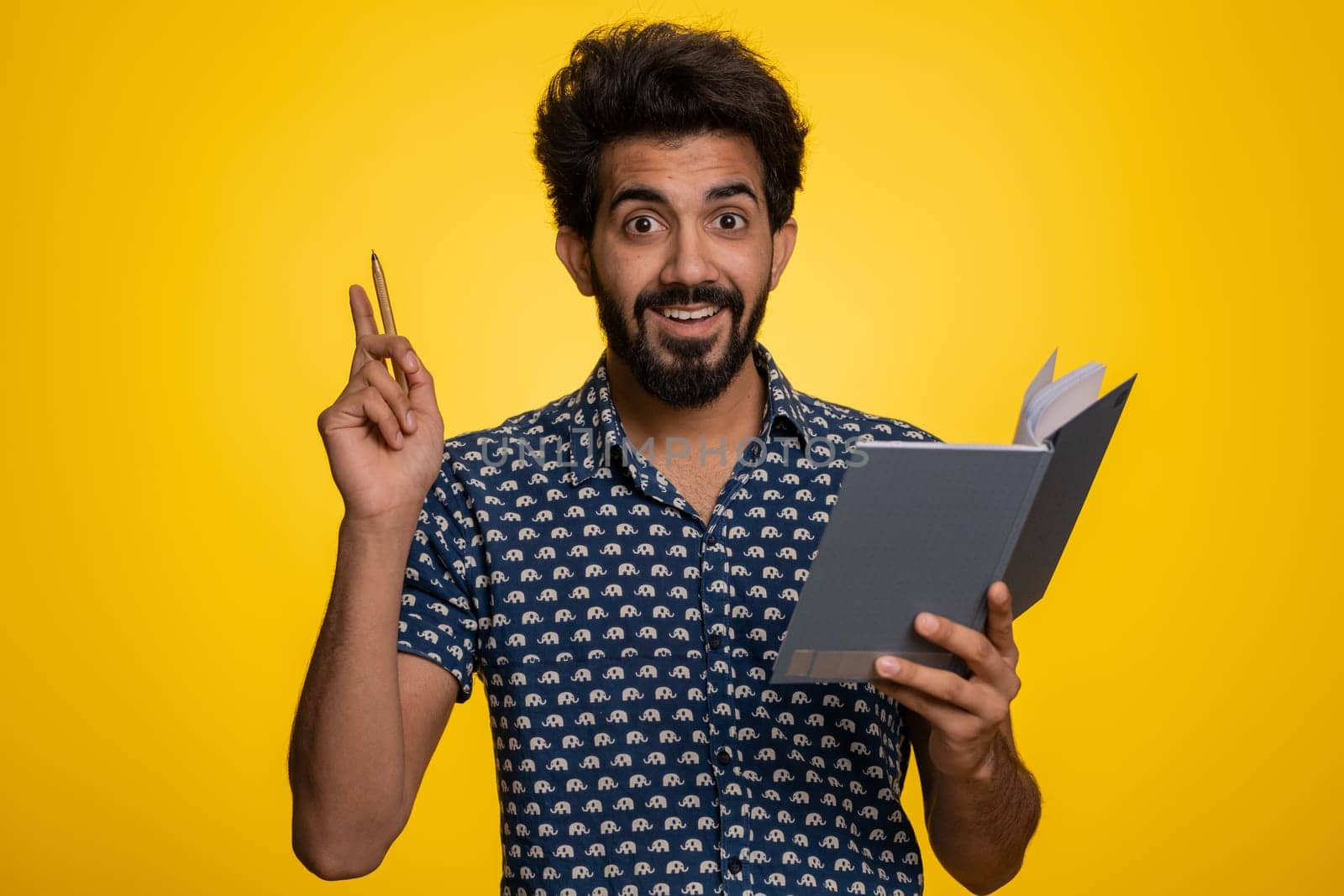 Thoughtful journalist indian man making notes, writing down thoughts with pen into notepad notebook diary, to do list, good idea. Handsome bearded hindu guy isolated alone on yellow studio background