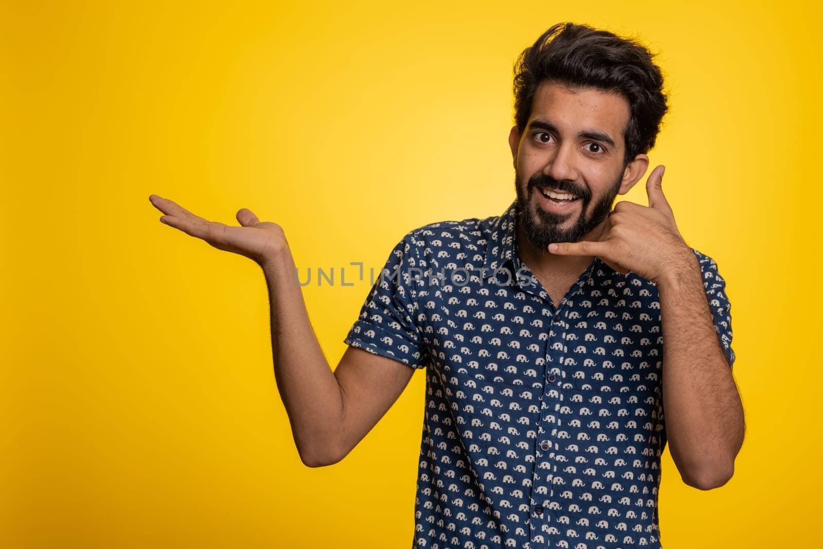 Call us, here is contact number. Handsome bearded indian man in shirt looking at camera doing phone gesture like says hey you call me back conversation. Hindu guy isolated on yellow background, indoor