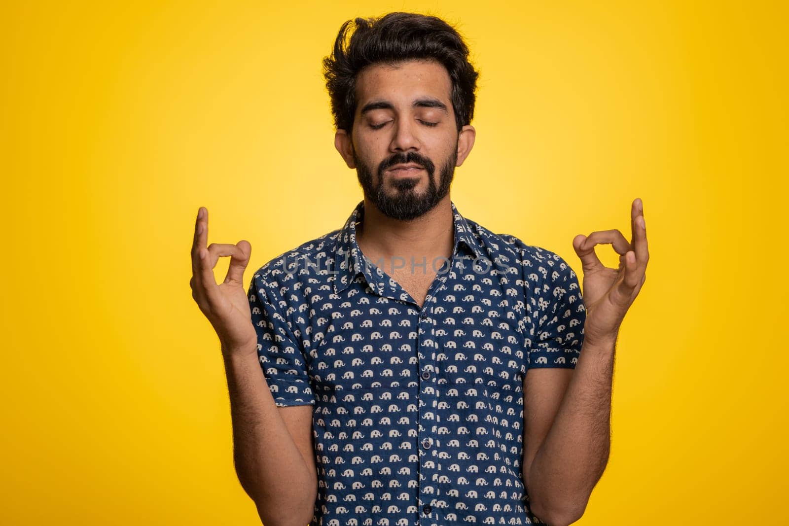 Keep calm down, relax, inner balance. Young indian man breathes deeply with mudra gesture, eyes closed, meditating with concentrated thoughts, peaceful mind. Hindu guy isolated on yellow background