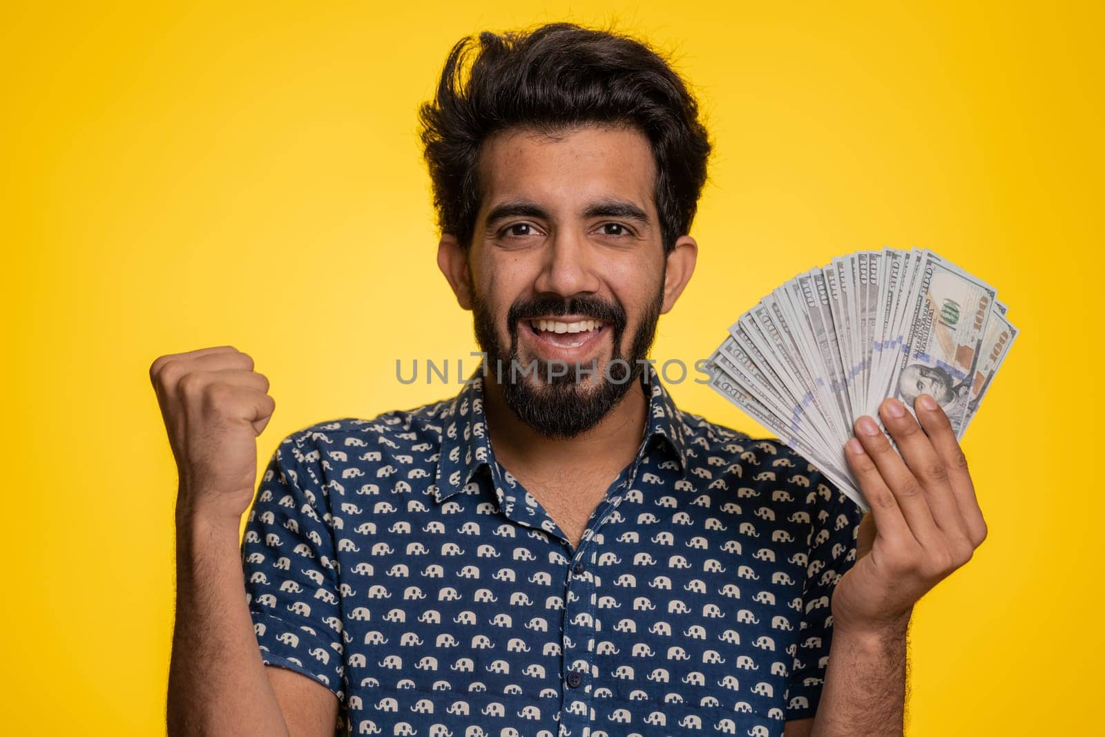 Rich pleased boss hindu man waving money dollar cash banknotes bills like a fan, success business career, lottery winner, big income, wealth. Handsome indian guy isolated on yellow background, indoor