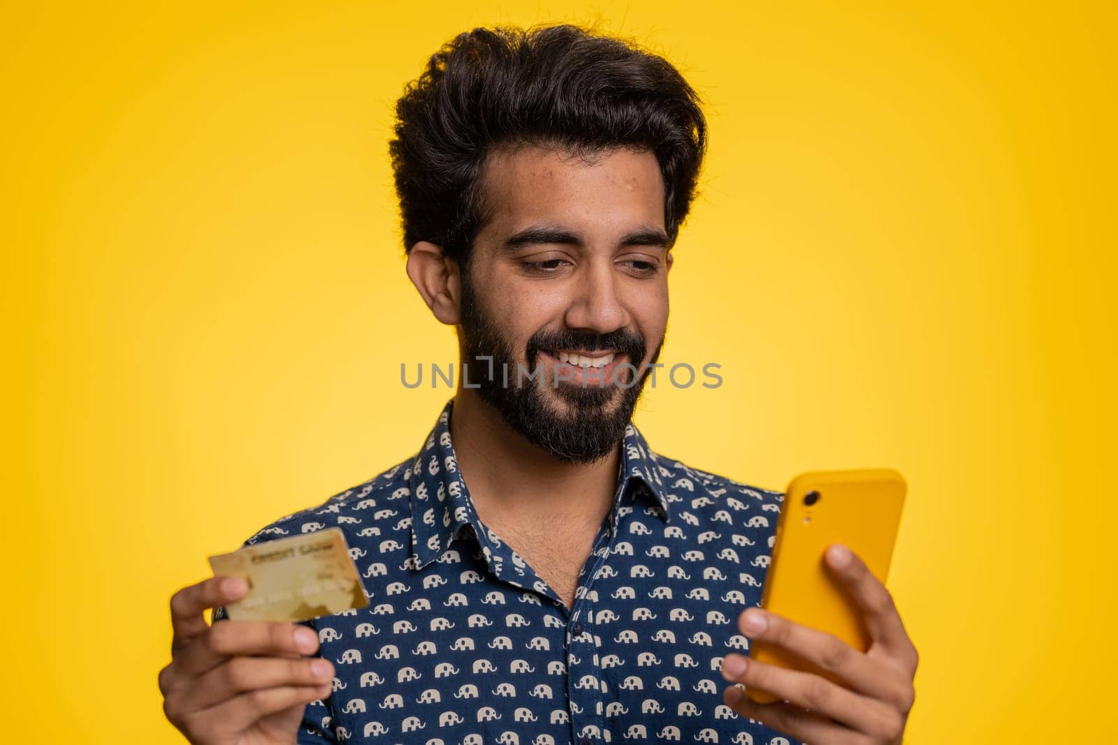 Sincere indian man customer using credit bank card and smartphone while transferring money, purchases online shopping, payment. Finance and internet. Handsome guy isolated alone on yellow background