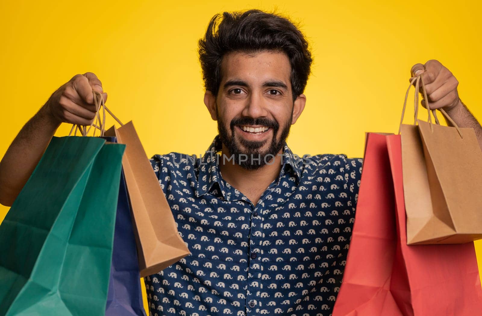 Young indian man showing shopping bags, advertising discounts, smiling looking amazed with low prices, shopping on Black Friday holidays. Shopaholic hindu guy isolated on studio yellow background