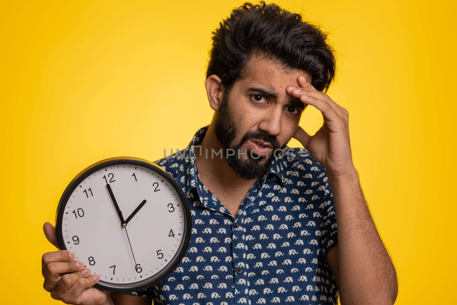 Young indian man with anxiety checking time on clock, running late to work, being in delay, deadline. Hindu guy looking at hour, minutes, worrying to be punctual isolated on studio yellow background