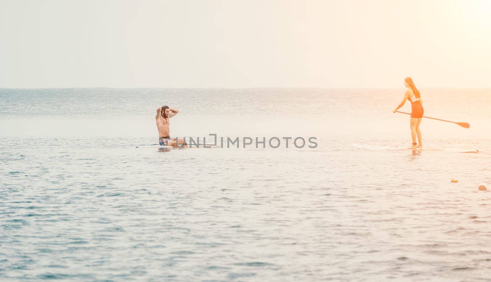 Sea woman and man on sup. Silhouette of happy young woman and man, surfing on SUP board, confident paddling through water surface. Idyllic sunset. Active lifestyle at sea or river. by panophotograph