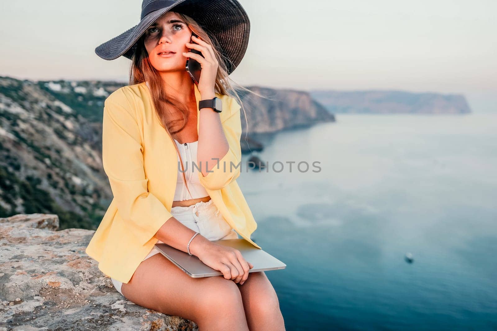 Successful business woman in yellow hat working on laptop by the sea. Pretty lady typing on computer at summer day outdoors. Freelance, travel and holidays concept.