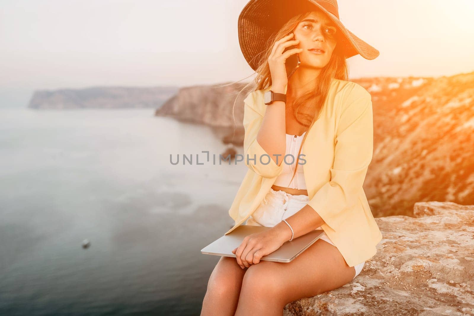 Successful business woman in yellow hat working on laptop by the sea. Pretty lady typing on computer at summer day outdoors. Freelance, travel and holidays concept.