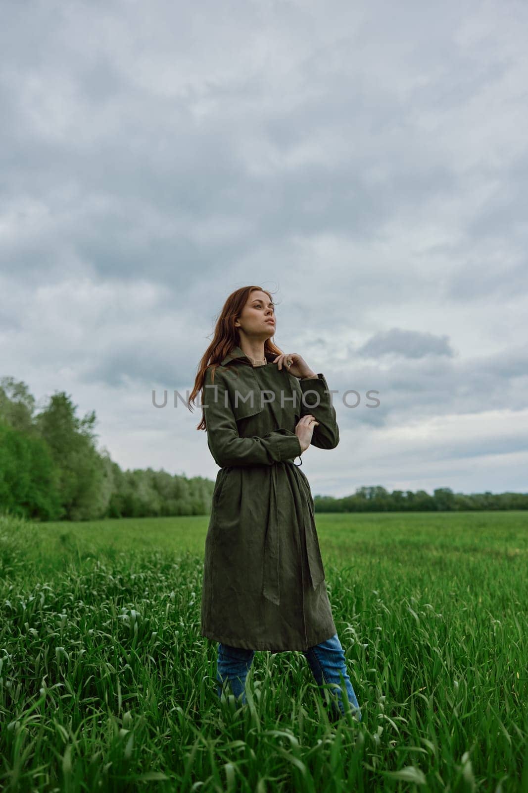 a beautiful woman in a dark coat stands in a green field in the spring in rainy weather. High quality photo