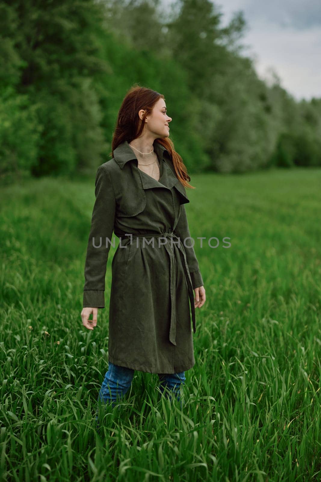 a woman in a long raincoat stands in tall green grass in a field in rainy weather in spring. High quality photo
