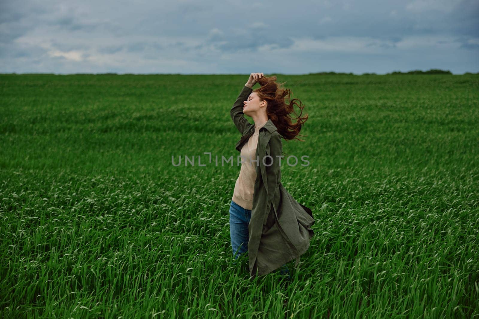 a woman in a dark coat stands in a green field against a cloudy sky holding her hair in the wind. High quality photo