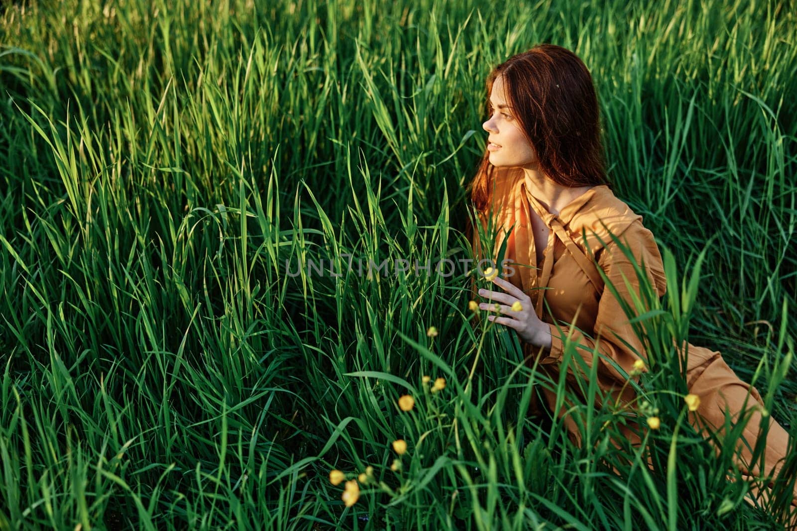 a happy woman is lying in the tall green grass in a long orange dress and smiling pleasantly looks away holding the leaves with her hand, enjoying nature. High quality photo