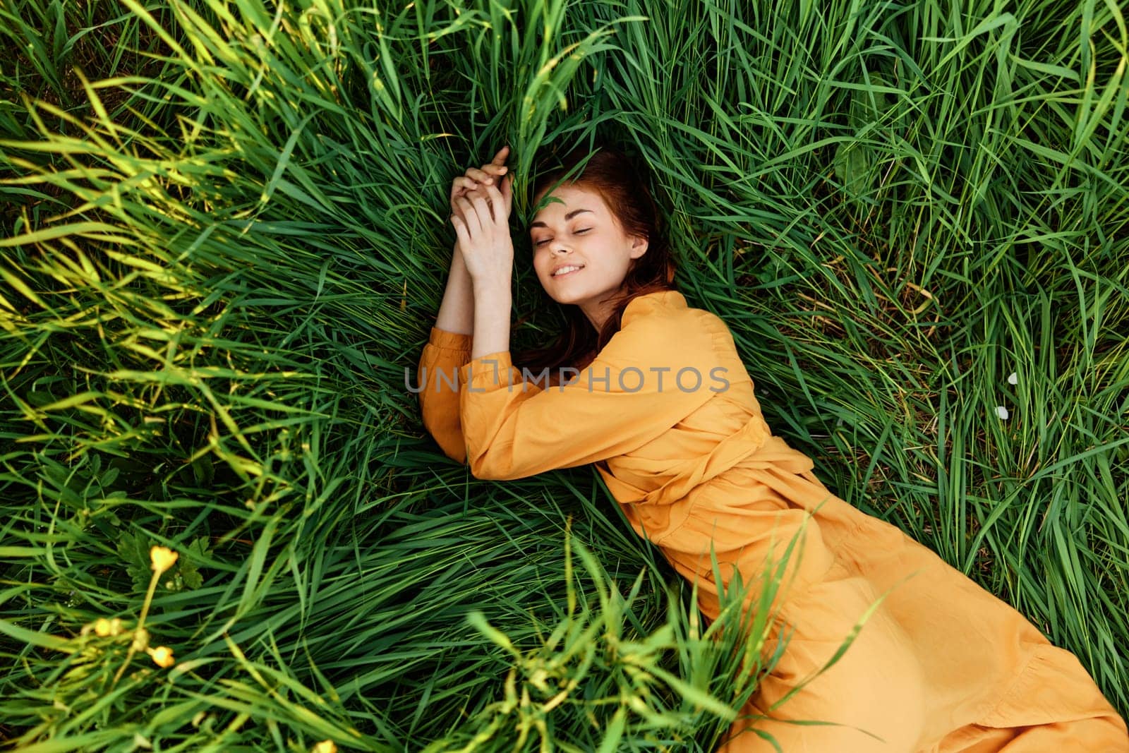 a relaxed red-haired woman enjoys summer lying in the tall green grass in a long orange dress smiling happily with her face turned to the camera by Vichizh