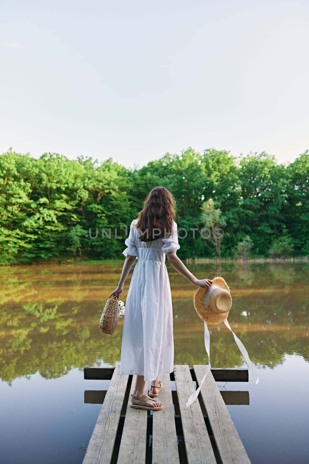 a woman in a long summer dress stands with her back to the camera on a pier near a lake in the forest with a wicker hat and a basket by Vichizh