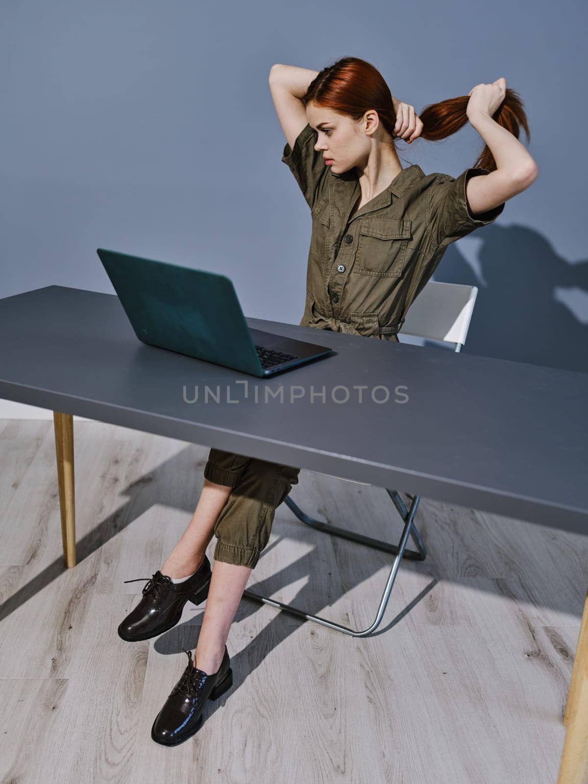 a woman in overalls sits at a laptop in the office and collects her hair with her hands. High quality photo