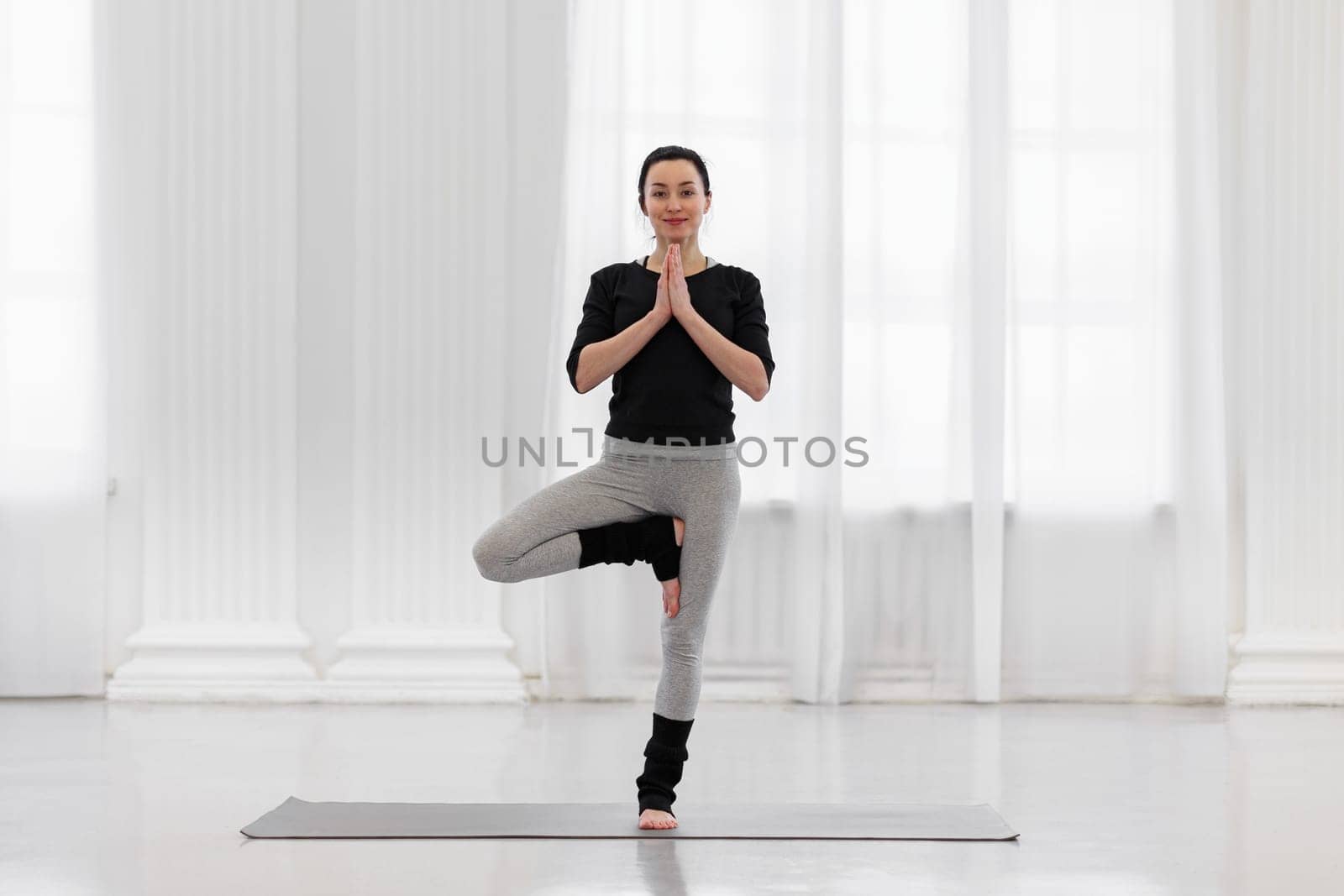 Pretty young woman doing vrikshasana on a mat on the floor in her spacious studio. Concept of benefits for the formation of correct posture heals, tones the muscles of the legs and spine by apavlin