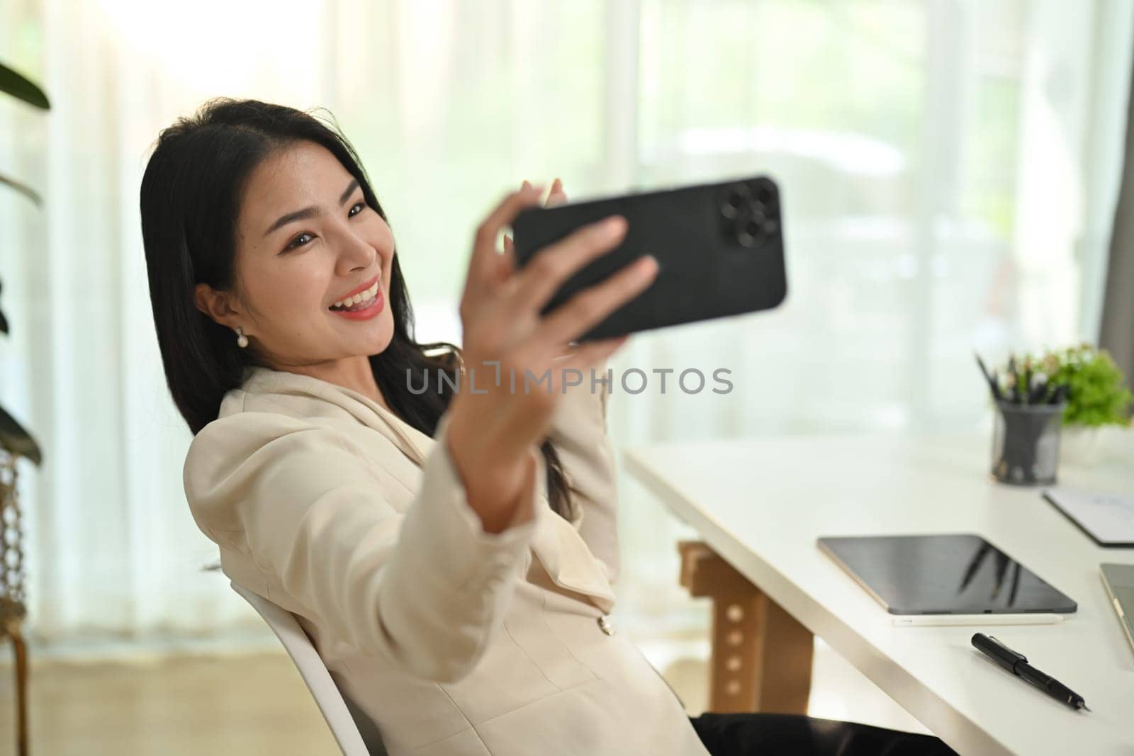 Cheerful young female entrepreneur making selfie or video call on her smart phone, sitting at working desk.