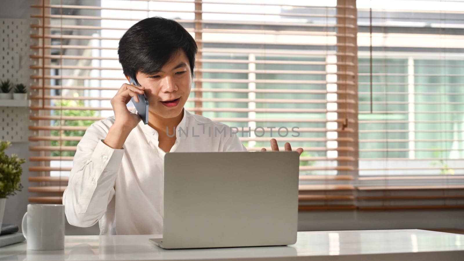 Confident male entrepreneur in white shirt having pleasant phone conversation and using laptop at home office.