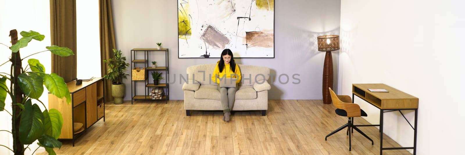 Young woman is working on laptop while sitting on couch in her comfortable living room. She is engaged in digital work, enjoying benefits of remote work and modern technology. High quality photo. by LipikStockMedia
