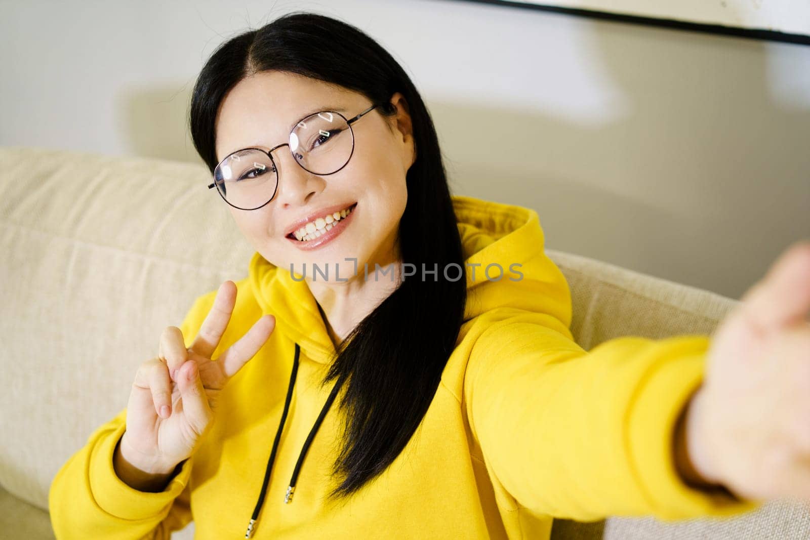 Asian woman takes self-portrait on her smartphone, smiling broadly and showing V symbol with her hand, expressing happiness and joy. High quality photo