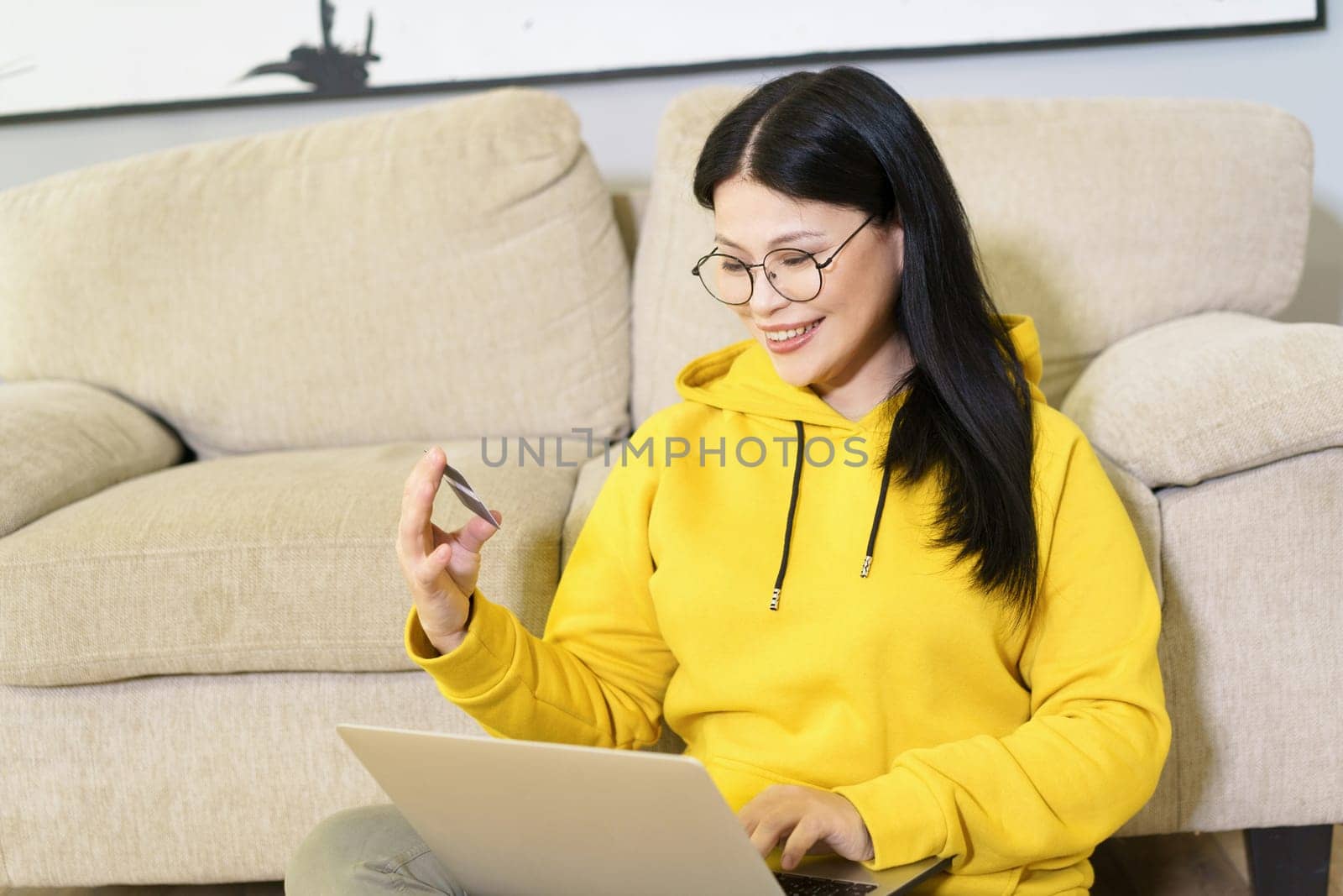 Asian woman comfortably shopping online from her home, with laptop on her lap and bank card in her hand. She is engaged in digital retail, using modern technology and the convenience of e-commerce to make purchase with her credit card. High quality photo