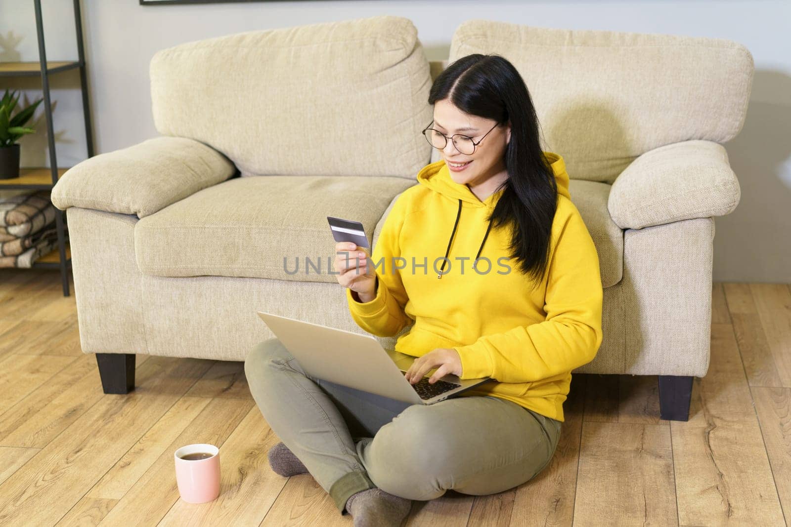 Mature Asian woman wearing glasses is sitting comfortably on floor with laptop, holding bankcard, and cup of coffee. She is engaged in digital work, enjoying moment of relaxation with her favorite beverage while working from home. High quality photo
