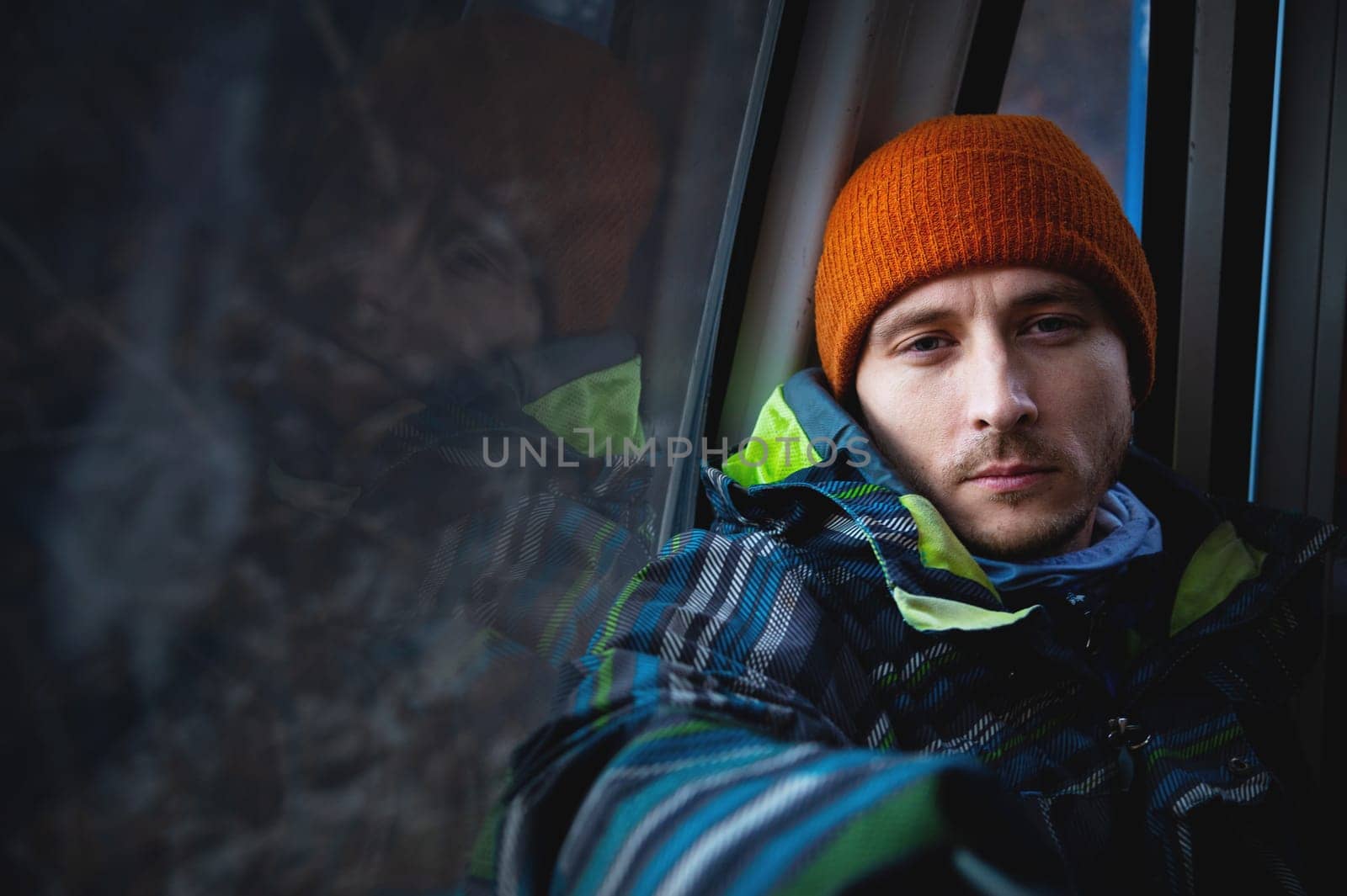 Portrait of a young man climbing to the top in the cabin of the cable car at a ski resort against the backdrop of mountains, on the side in the mirror his reflection by yanik88