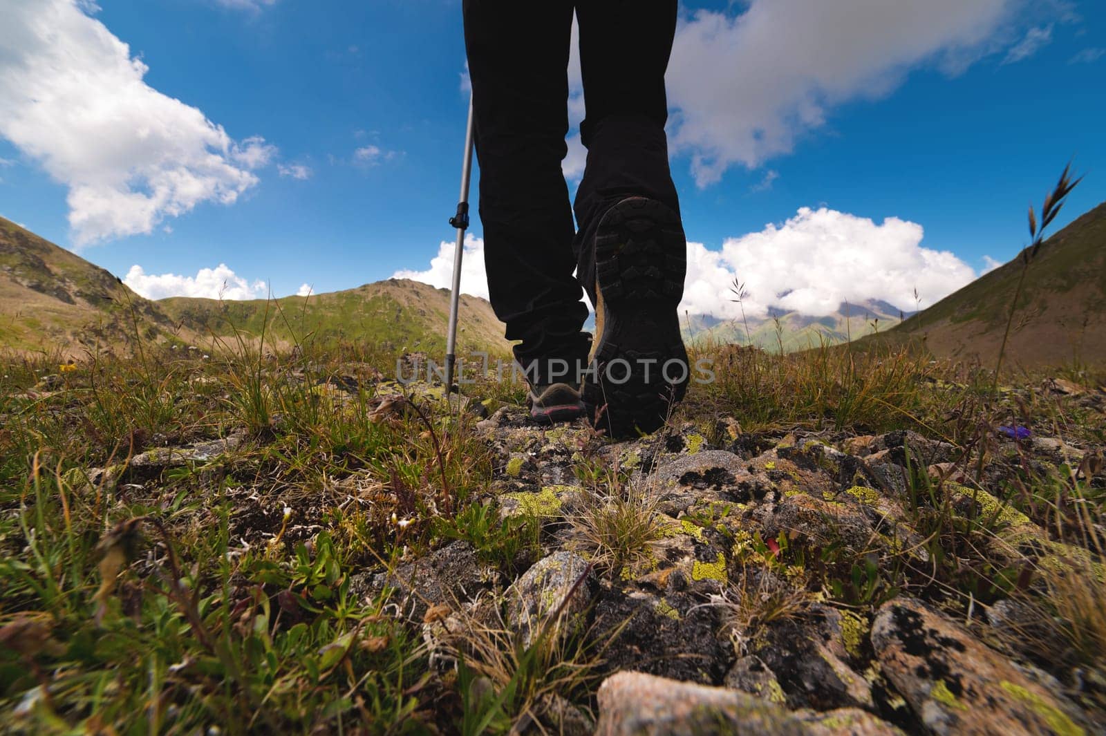 Hiking trail with flowers, green grass and stones. Close up of hiking boots in the mountains against the backdrop of mountains and fluffy clouds.