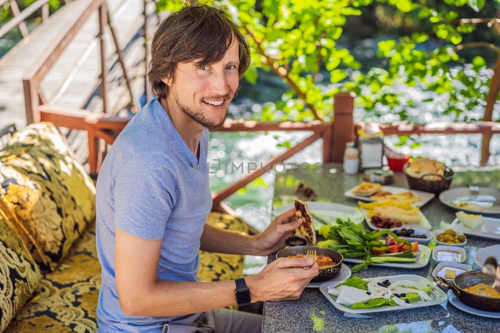 man tourist eat Turkish breakfast. Turkish breakfast table. Pastries. Vegetables. Olives. Cheeses, fried eggs. Jams, tea in copper pot and tulip glasses. Wide composition.