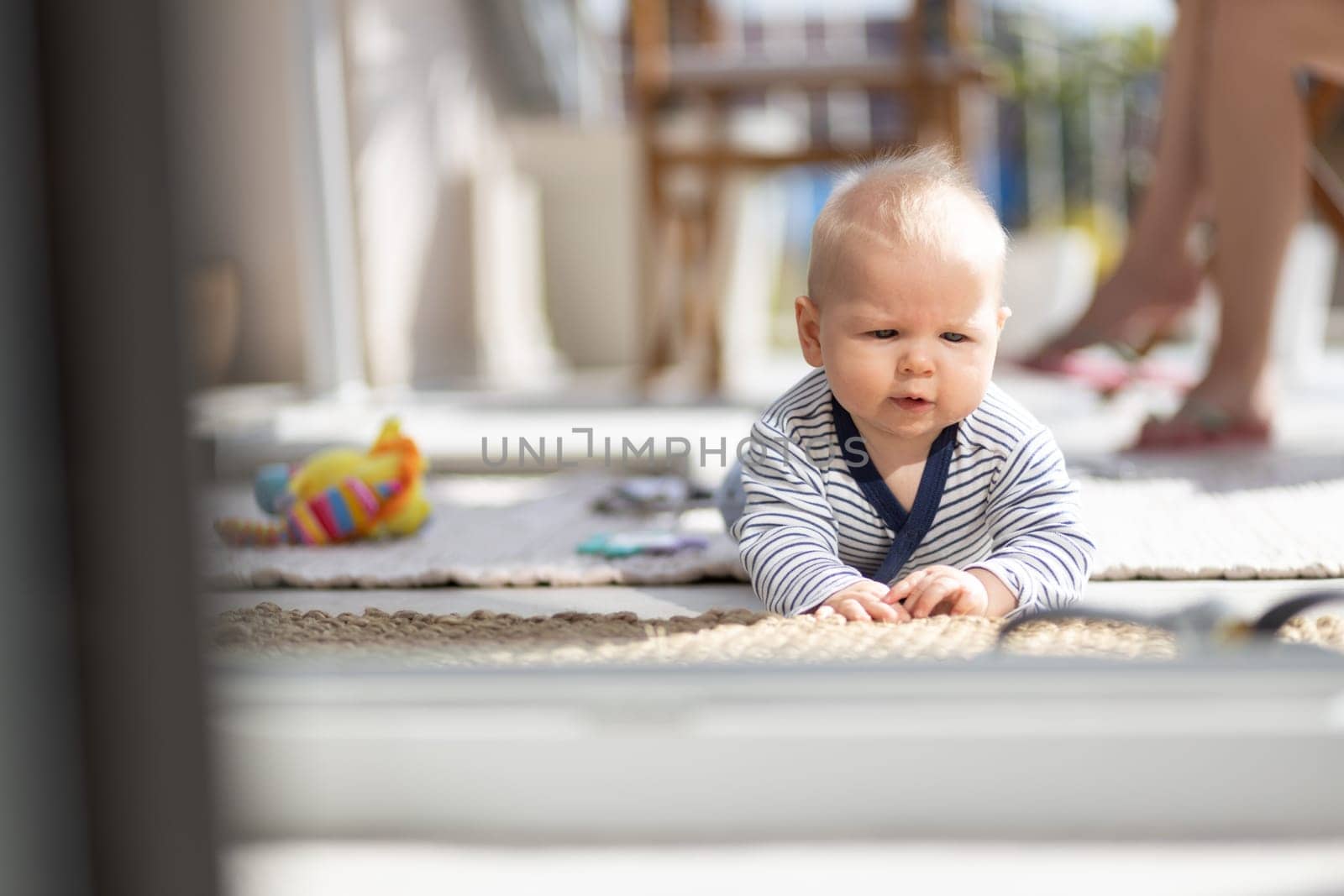 Cute little infant baby boy playing with toys outdoors at the patio in summer being supervised by her mother seen in the background. Selective focus.