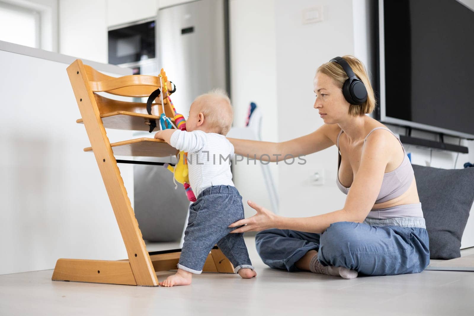 Women's multitasking. Mother sitting on floor playing with her baby boy watching and suppervising his first steps while listening to podcast on wireless headphones