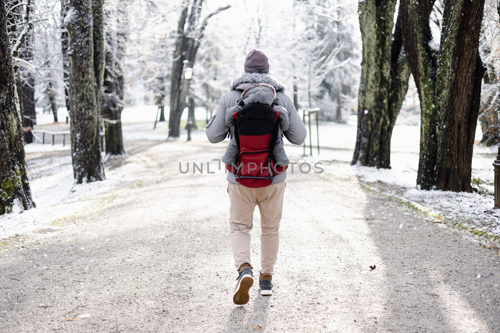Rear view of sporty father carrying his infant son wearing winter jumpsuit and cap in backpack carrier walking in city park in winter.