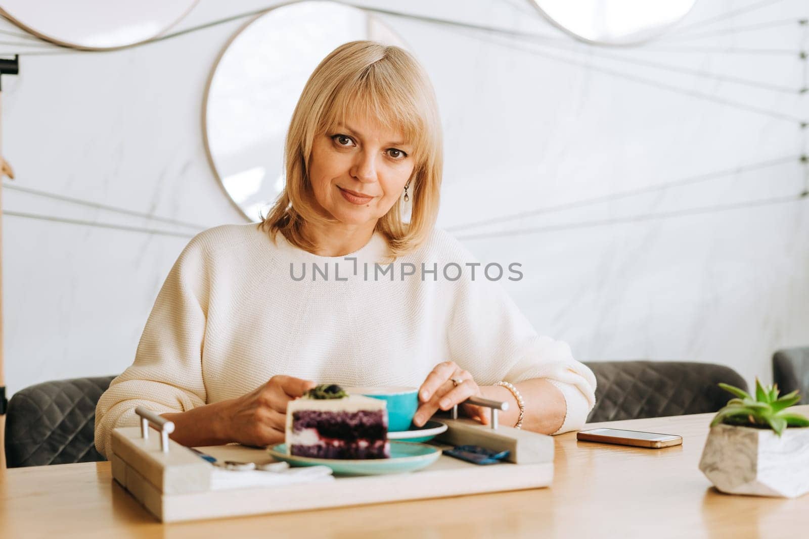 Mature woman holding a cup of cappuccino coffee and a cream cake. Adult lady in white sweater enjoying her breakfast latte and dessert pie on a wooden tray in a cafe.