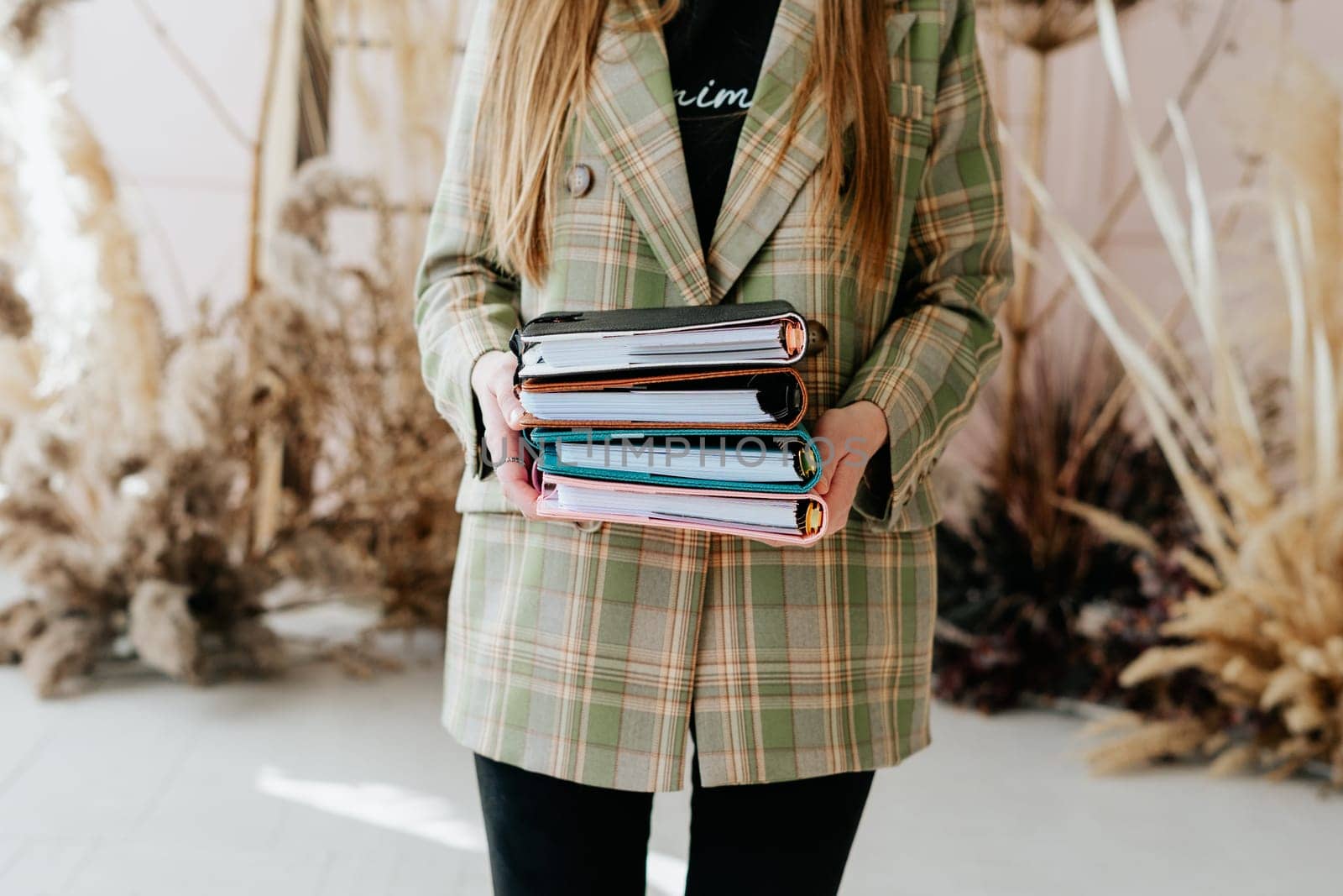 Business woman office secretary suit holding stack of notebooks. Student with a stack of notepads and dry flowers in the background. Cropped girl in a stylish office outfit holds books in her hands.
