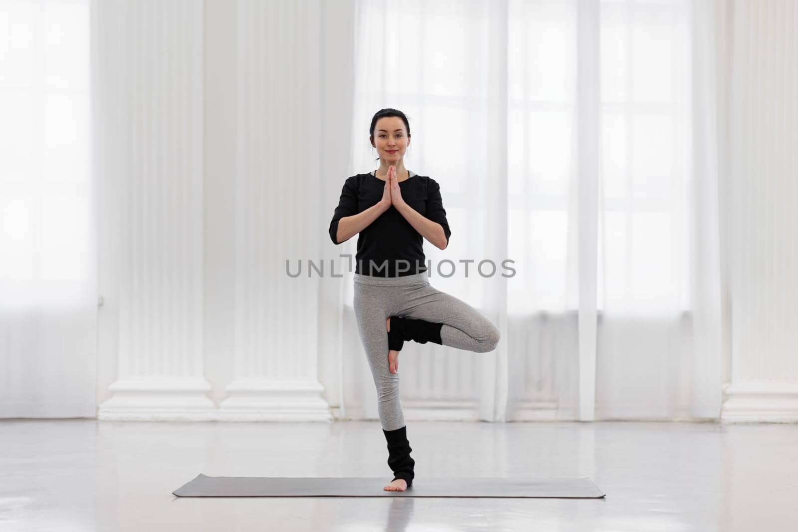Beautiful young woman doing tree pose during yoga meditation in the white gym. The concept of awareness, calmness of the nervous system, balance and harmony with oneself by apavlin