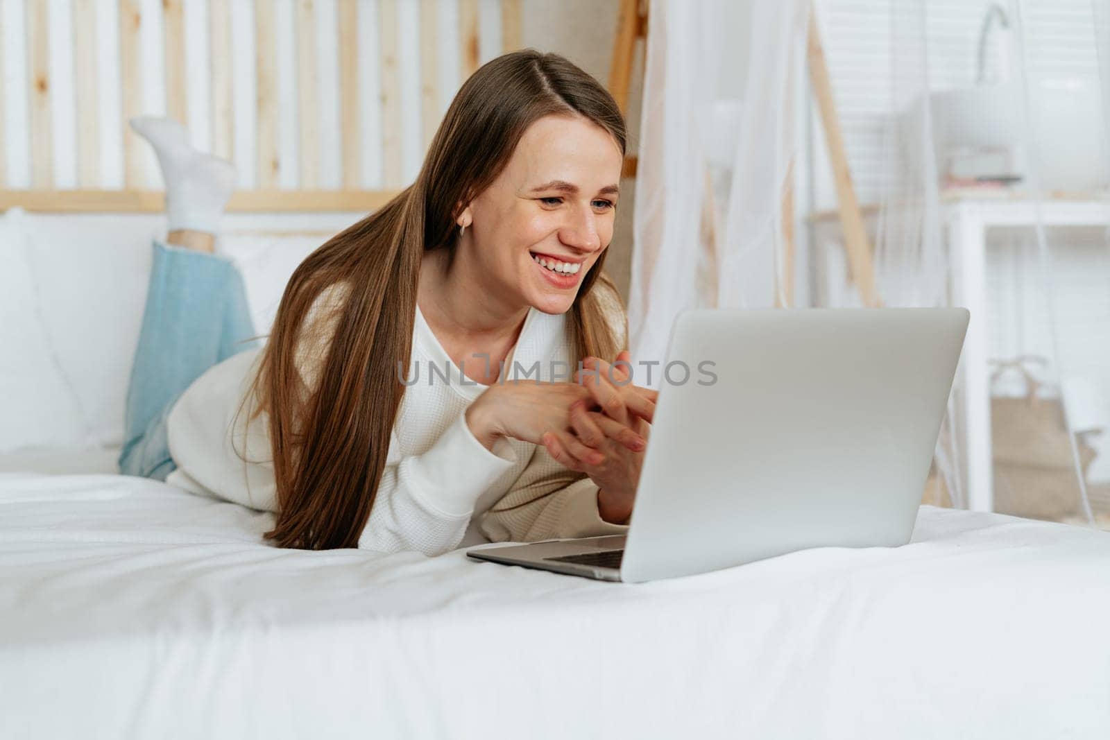 Freelance woman having a video call chat, typing in laptop and online shopping, lying on the white bed. Happy relaxed girl woking from home office. Distance learning online education and work.