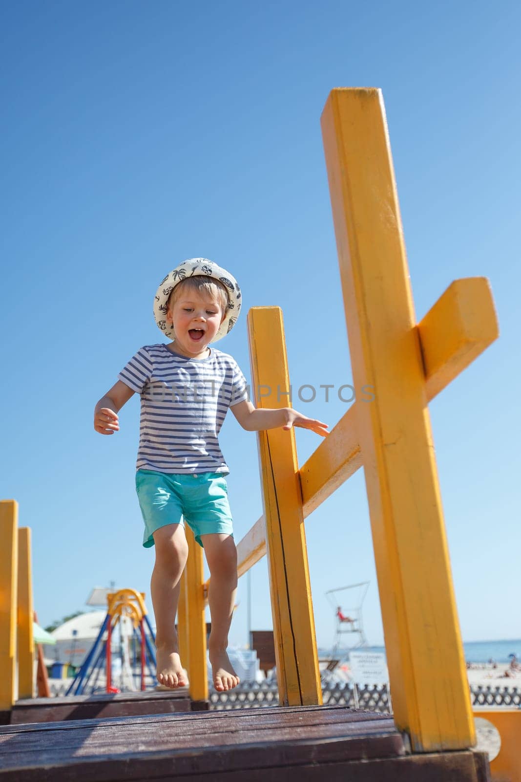 A little boy with a hat and a striped sailor shirt play in the playground jumping on a wooden bridge