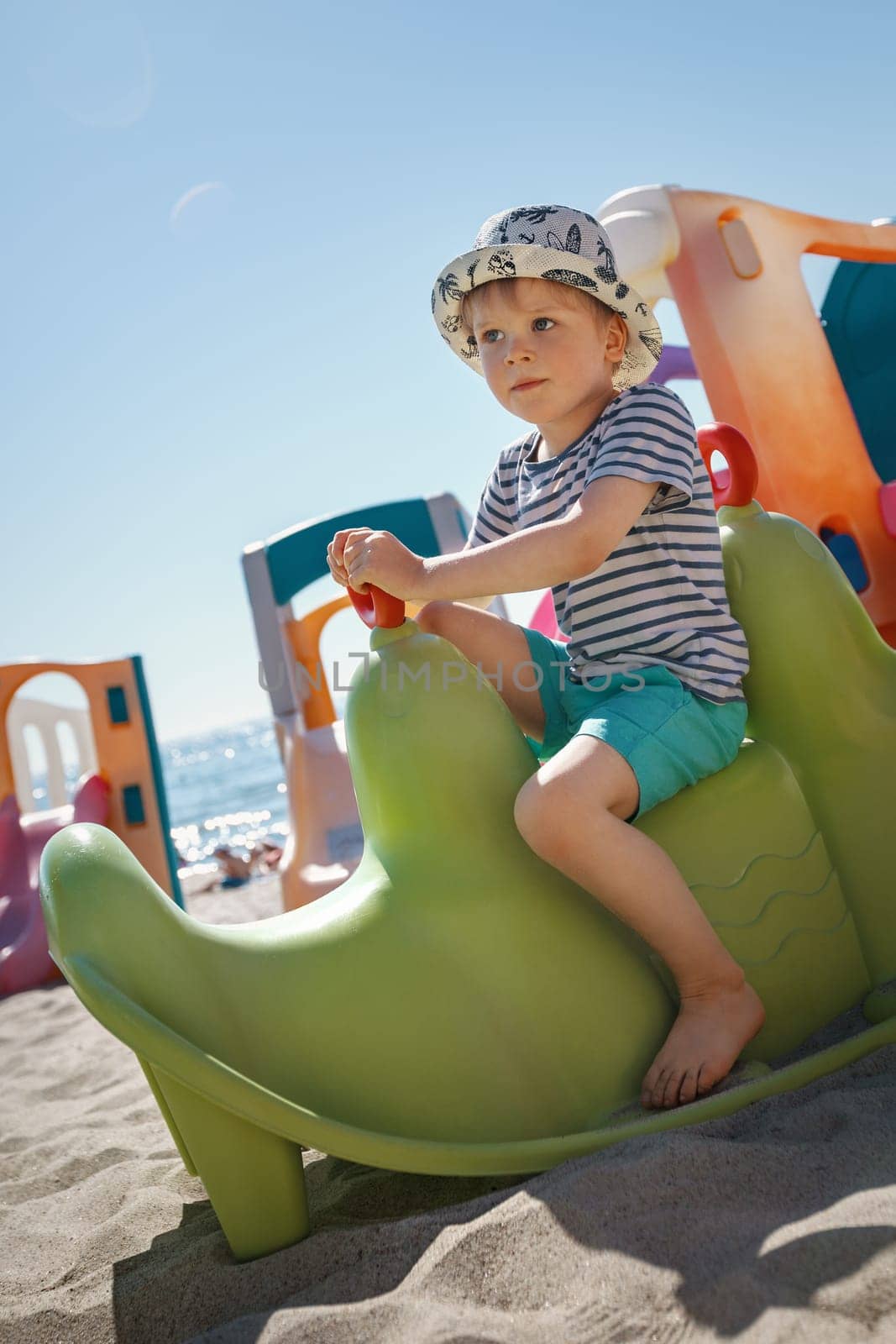 A little boy poses on a green plastic toy at a beach playground. Sand, sea and light blue sky in the background. by Lincikas