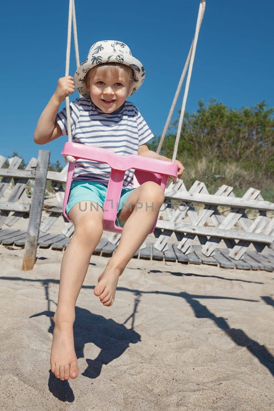 The little hilarious boy swings in a pink swing, he looks straight at the camera and poses. Beach background, sand and blue sky