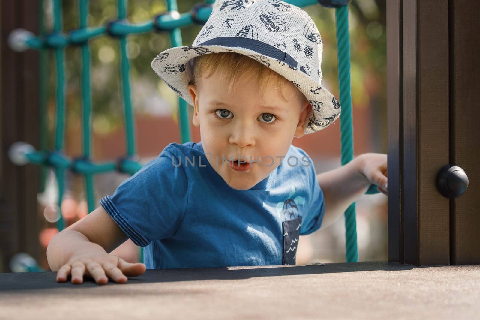 Cute little boy playing on the playground in the summer. A portrait of a child in a hat looks straight into the camera with a surprised facial expression.