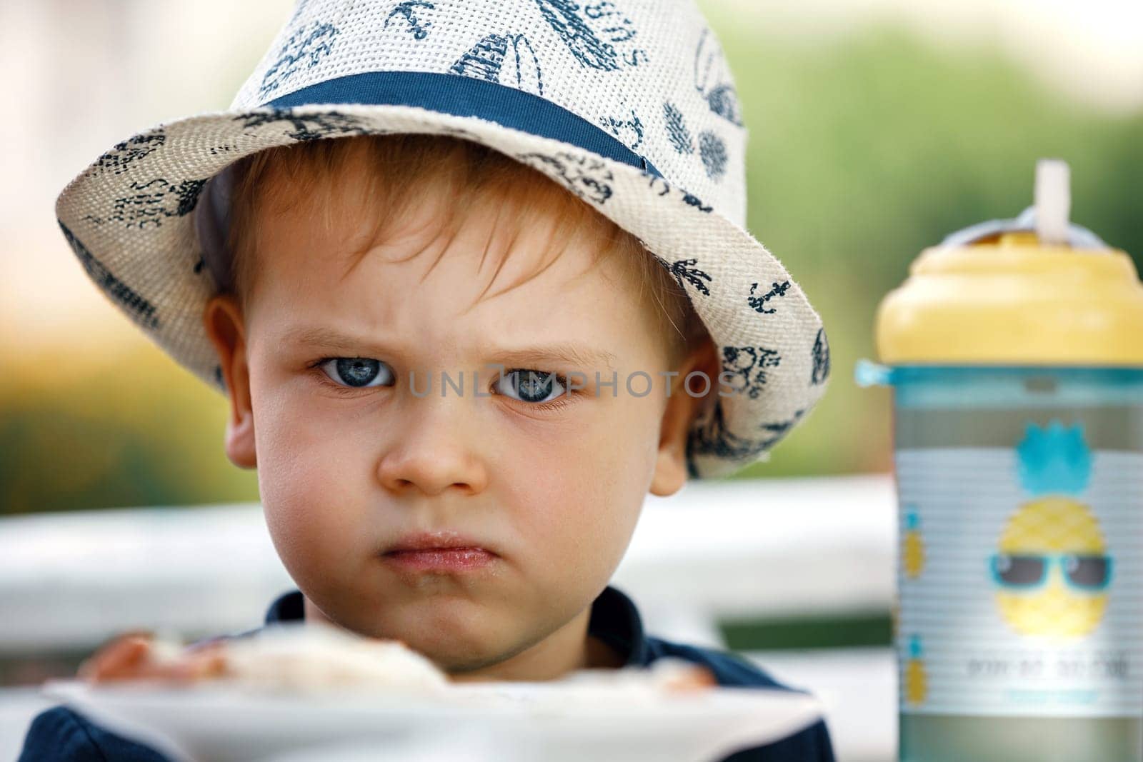 Close up portrait of small angry caucasian boy in hat unwilling to eat food. by Lincikas