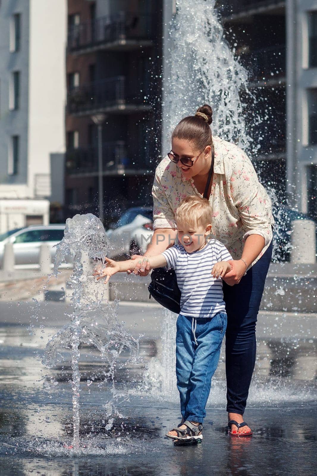 Mom and her son, in the city center, by a fountain of water. The child is afraid to touch the water so that it does not get wet, the mother encourages it by Lincikas