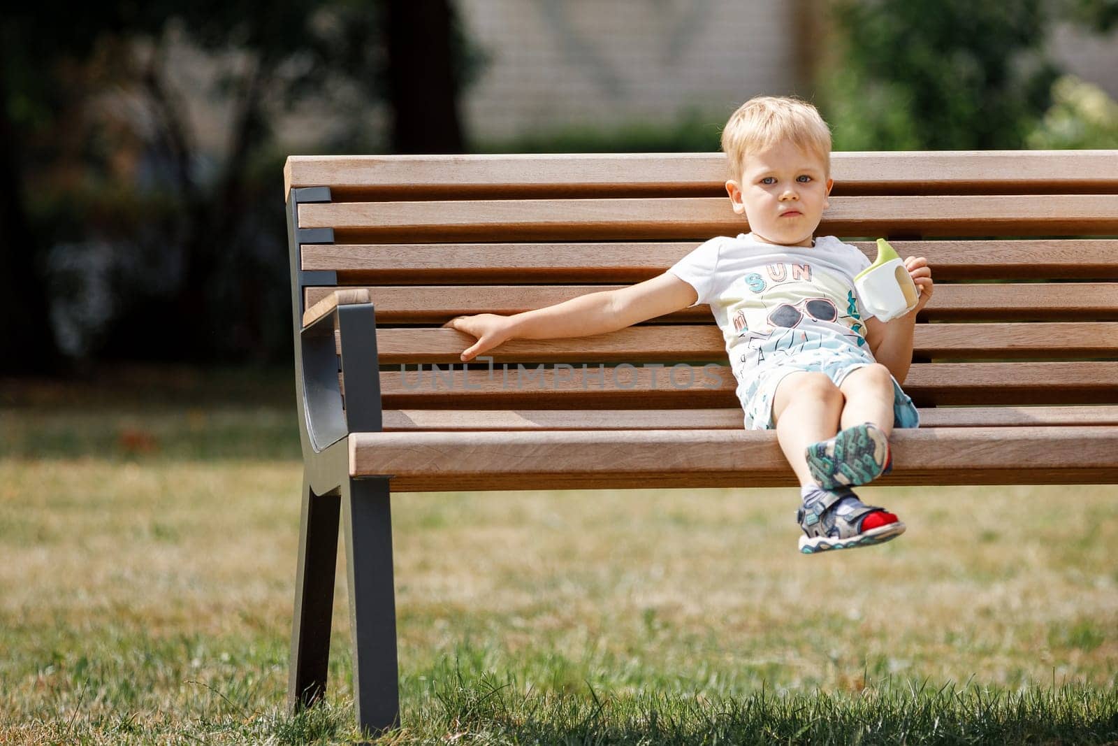 Little boy with is sitting on bench in park and drinks juice from plastic cup. by Lincikas