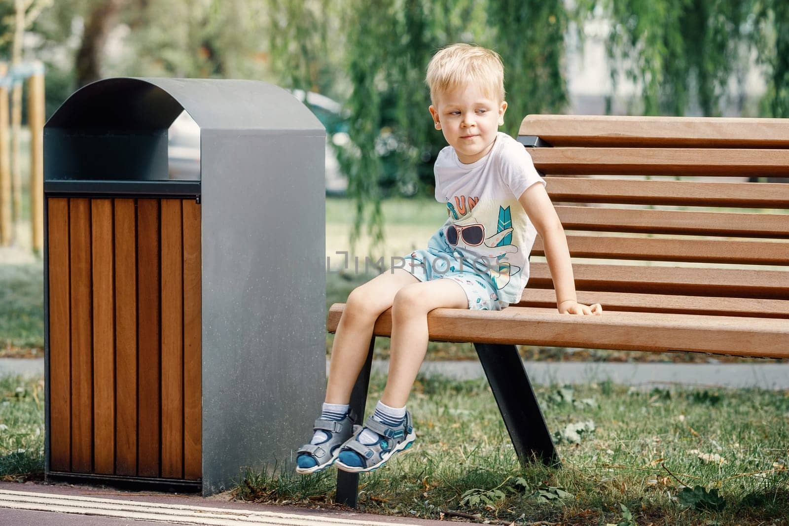 Child portrait sitting on a bench in park, white haired boy 3 years old. Beautiful bench and trash can with a modern design.
