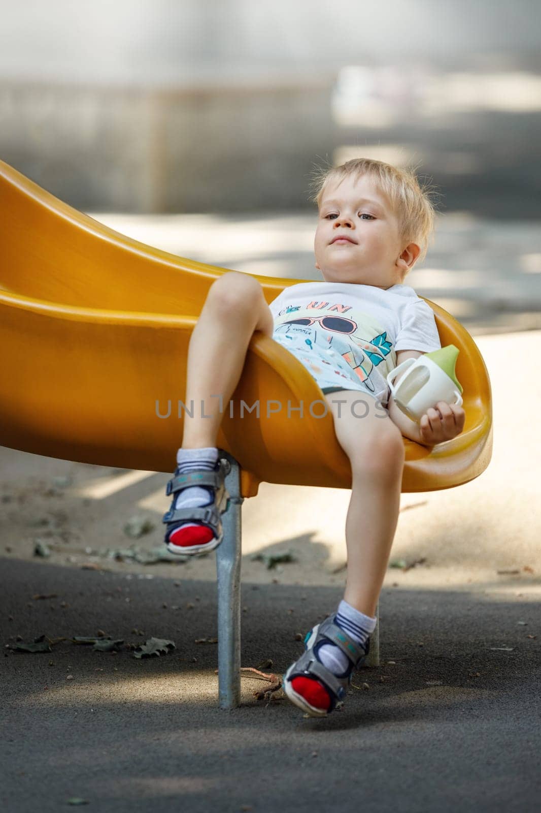 The little boy slips into a yellow plastic slide, his hair becomes eclectic, child sits down comfortably to calm and drink. by Lincikas