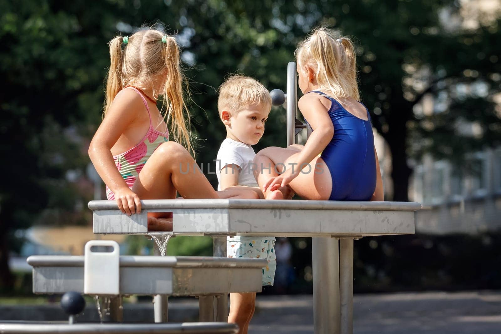 A happy boy and a cute girls in a blue bathing suit play with a water tap in a city park. by Lincikas