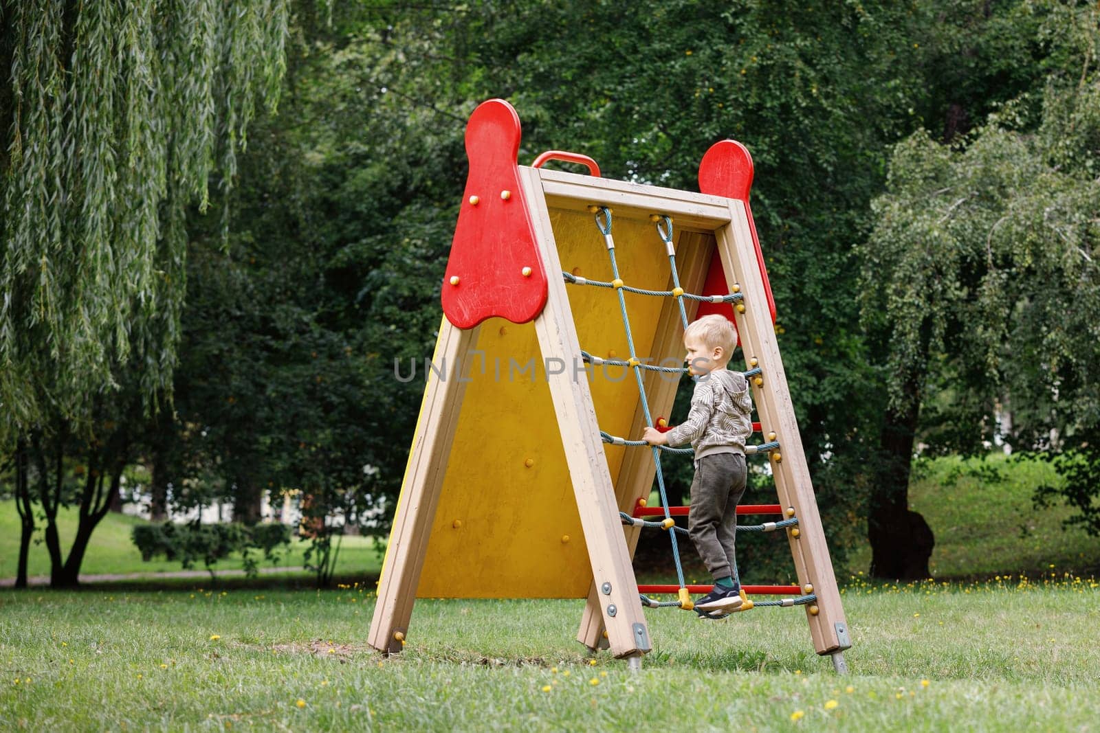 Little boy climbs in a red rope wall web on the playground in the park.