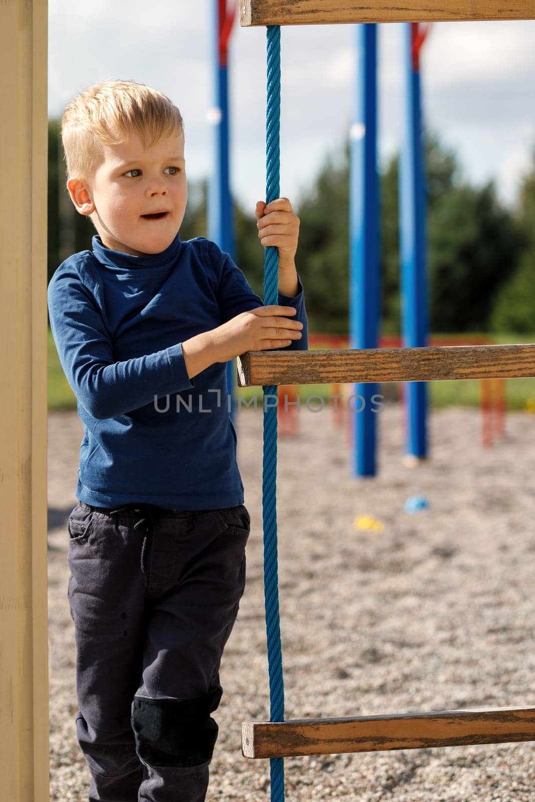 An active child in a blue sweatshirt on a playground next to a rope ladder by Lincikas