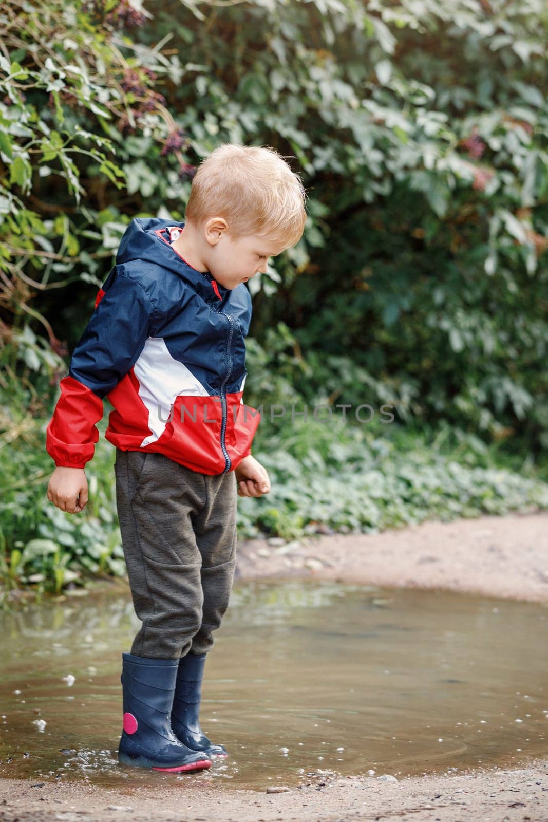 Little boy in colourful raincoat and rubber blue boots playing in puddle.