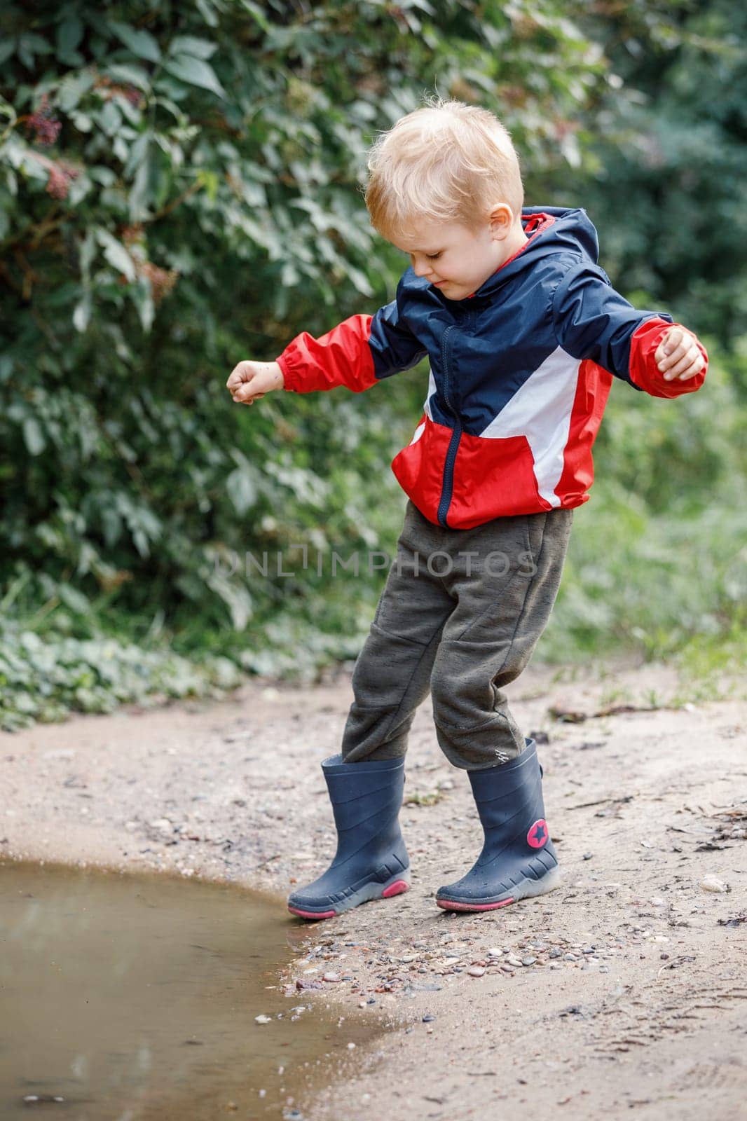 A timid little boy from the city in a red jacket, carefully trying to go the puddle.