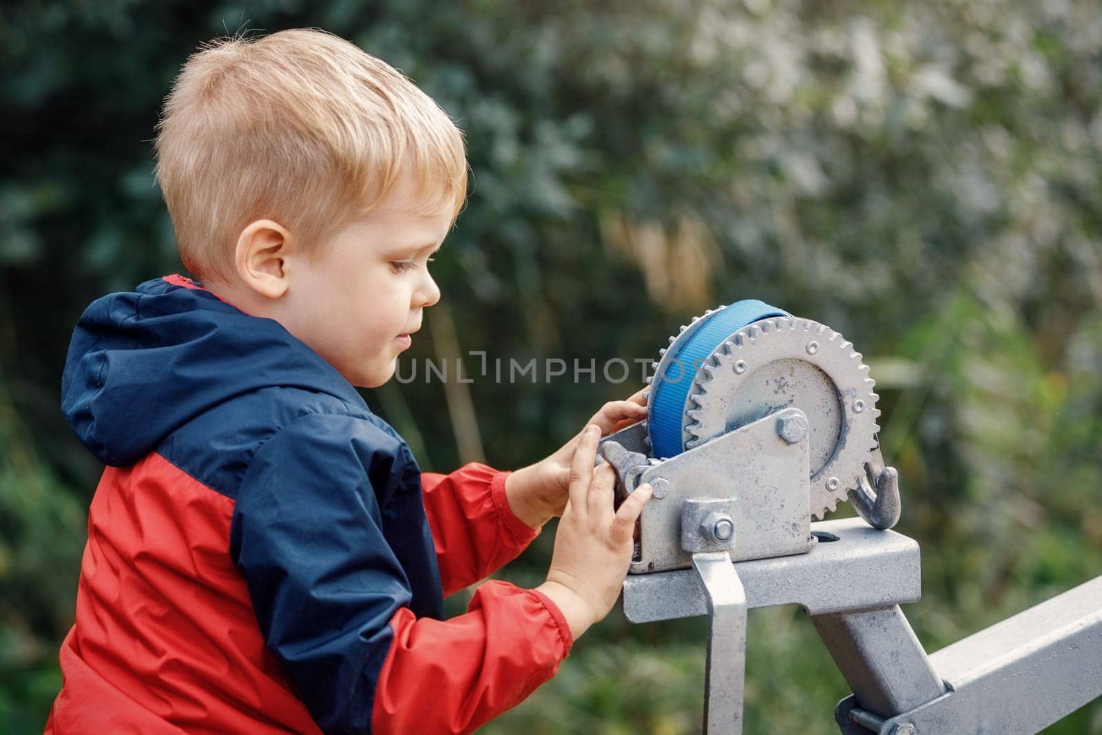 The little boy is interested in playing with boat trailer winch, boat bow rest, blue cargo strap and its pulley and gears.