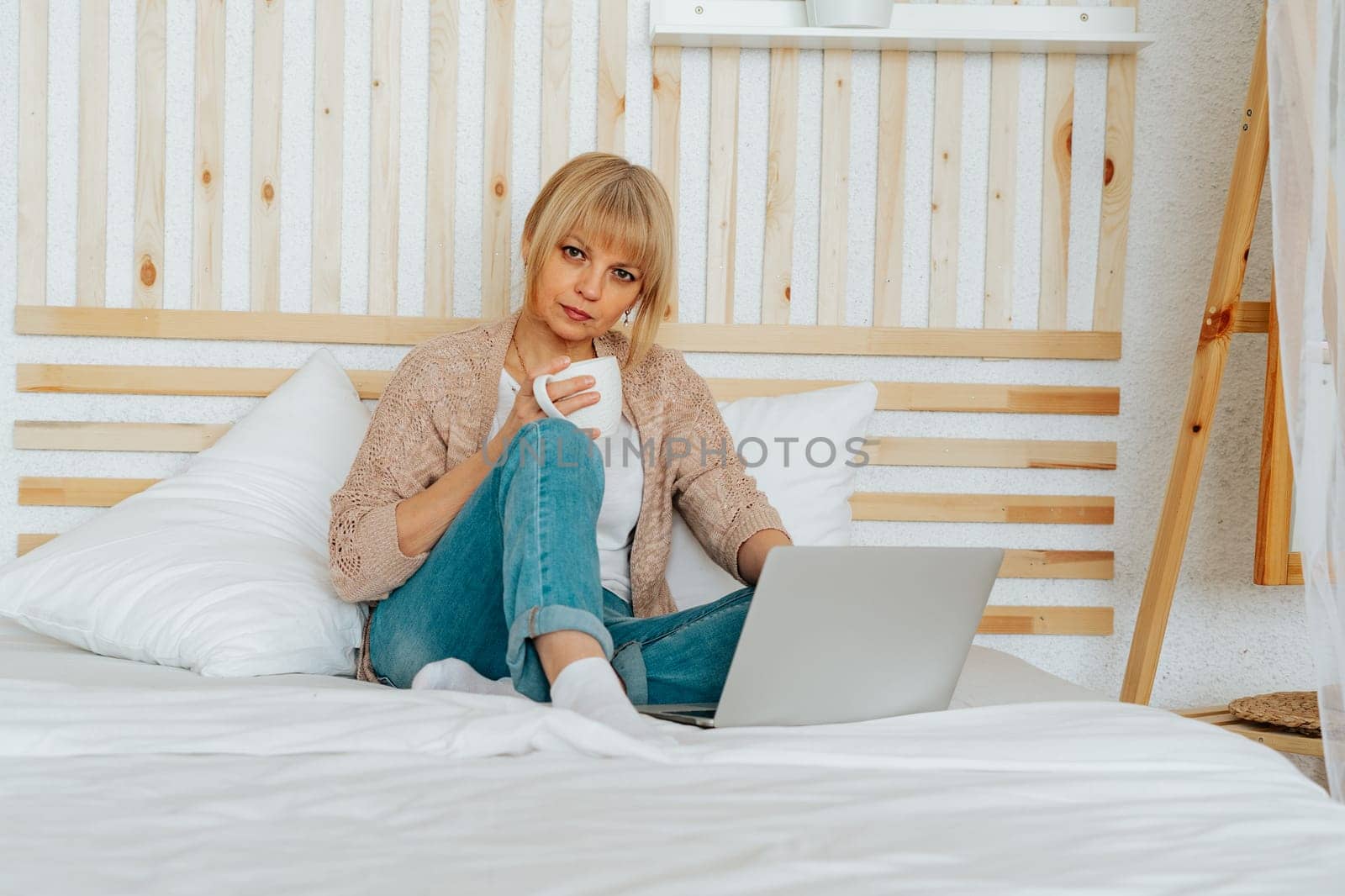 Distance learning online education and work. Relax mature woman typing at laptop, sitting on the bed and drinking coffee. Smiling senior with notebook at home office. Using computer and shopping.